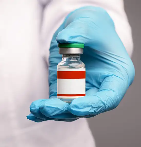 A hand in a blue medical glove holds a small glass vial with a green cap and a blank red-and-white label.