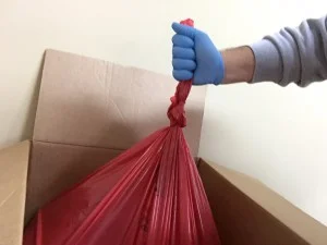 Red Bag Setup A hand wearing a blue medical glove lifting a red plastic trash bag out of a cardboard box.