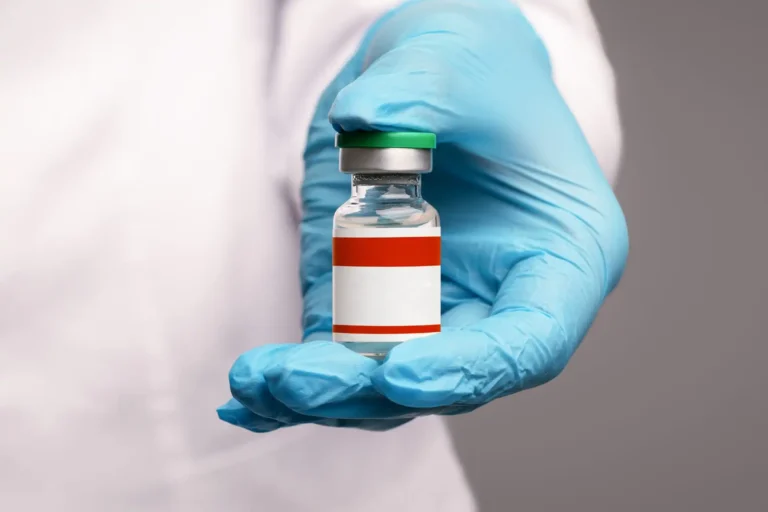 A person wearing a blue medical glove holds a small glass vial with a green cap and a blank red-and-white label.