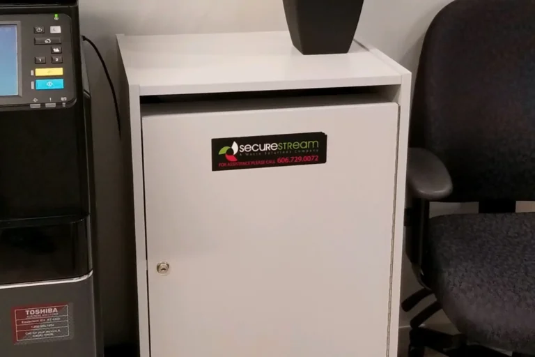 A white secure document disposal bin with a SecureStream logo and phone number, positioned next to a printer and a desk chair.