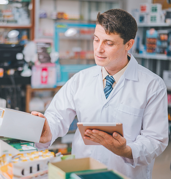 A pharmacist in a white lab coat and tie holds a tablet while checking a product box in a pharmacy.